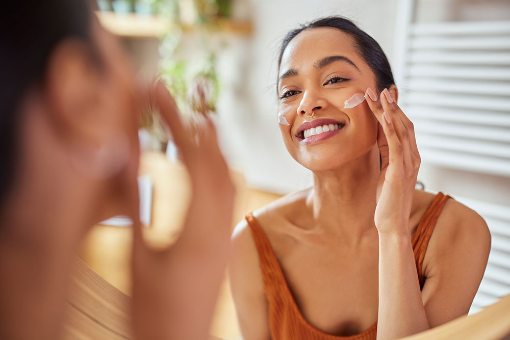 Portrait of smiling latin woman applying cream on her face while looking in mirror in her bathroom. Young multiethnic woman applying moisturizer on her cheek while looking in the mirror.