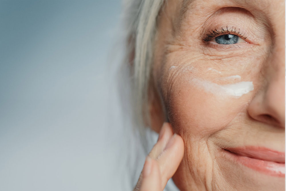 Portrait of smiling latin woman applying cream on her face while looking in mirror in her bathroom. Young multiethnic woman applying moisturizer on her cheek while looking in the mirror.